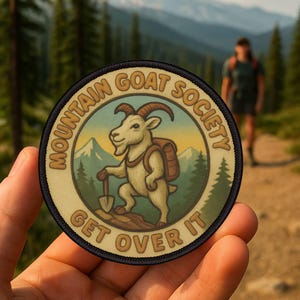 Può includere: Una toppa rotonda con la scritta "MOUNTAIN GOAT SOCIETY" e "GET OVER IT". Il design presenta un cartone animato di una capra con uno zaino e una pala, sullo sfondo di una montagna e di una foresta. La toppa è tenuta in mano.