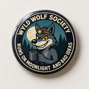May include: A circular pinback button featuring a cartoon wolf wearing a headlamp, holding a coffee mug, set against a forest and moon background. The text reads WYLD WOLF SOCIETY RUNS ON MOONLIGHT AND BAD IDEAS in a circular border.