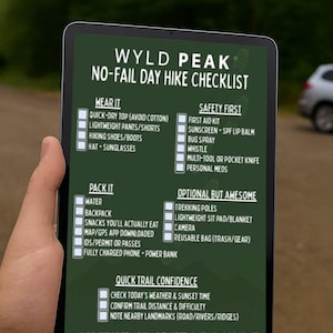 Pode incluir: Um tablet exibe uma lista de verificação de caminhada de um dia "WYLD PEAK". A lista inclui itens para usar, embalar, itens de segurança e equipamentos opcionais. O fundo é desfocado.