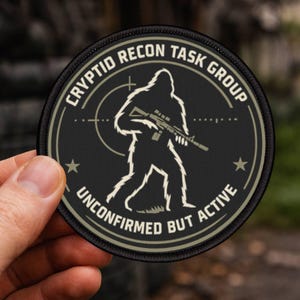 May include: A circular black patch featuring the text "CRYPTID RECON TASK GROUP" and "UNCONFIRMED BUT ACTIVE". The design includes a white Bigfoot silhouette holding a rifle, set against a target. The patch has a tan border and two stars.