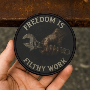 Puede incluir: Parche redondo negro con el texto "FREEDOM IS FILTHY WORK" en blanco. El parche presenta una ilustración detallada de una mano agarrando una llave. La llave es plateada y la mano es una ilustración detallada.
