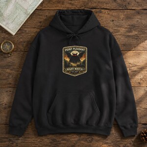 May include: A black hooded sweatshirt featuring a graphic print of a winged creature with glowing red eyes above a forest silhouette. The text reads Point Pleasant Night Watch Est. 1967. The hoodie includes a front kangaroo pocket and drawstrings.