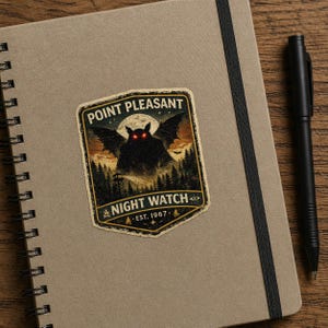 May include: A tan notebook with a black elastic closure and a sticker featuring the text "Point Pleasant Night Watch Est. 1967" and a graphic of a winged creature with red eyes. A black pen rests on the right side.