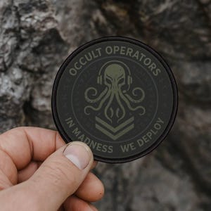 May include: A round, black patch with the words "OCCULT OPERATORS" and "IN MADNESS WE DEPLOY" encircling a central design. The design features an octopus wearing headphones. The patch is held in a hand.