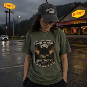May include: An olive green t-shirt with a Point Pleasant Night Watch graphic featuring a black creature with red eyes, a full moon, and forest scene. The person is wearing a grey West Virginia baseball cap.