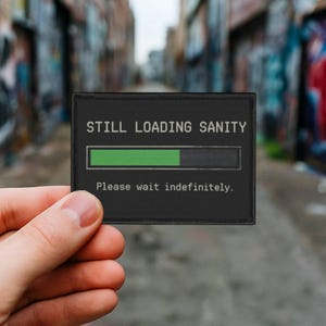 Könnte beinhalten: Ein schwarzer rechteckiger Aufnäher mit dem Text "STILL LOADING SANITY" über einem grünen Fortschrittsbalken. Darunter steht "Please wait indefinitely."