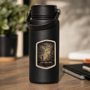 May include: A black insulated water bottle with a black handle. A sticker on the bottle features a vintage map design with the text "LAST SEEN HERE." The bottle is on a wooden surface.