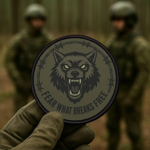 Peut inclure: Un écusson rond vert olive avec une tête de loup menaçante entourée de fil de fer barbelé. Le texte "FEAR WHAT BREAKS FREE" est inscrit en arc de cercle autour du loup. L'écusson est tenu par une main gantée, avec des silhouettes floues en tenue militaire en arrière-plan.