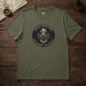 May include: An olive green short-sleeve t-shirt featuring a circular graphic on the chest. The design displays a stylized octopus wearing headphones above two chevron stripes, surrounded by the text "OCCULT OPERATORS" and "IN MADNESS WE DEPLOY".