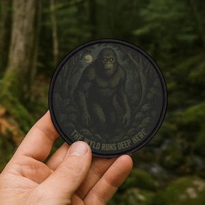 Può includere: Toppa rotonda nera con un'illustrazione scura di una creatura simile a Bigfoot in una foresta sotto la luna piena. Il testo "THE WYLD RUNS DEEP HERE" è stampato attorno al bordo della toppa.