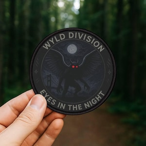 May include: A round, black patch with the text "WYLD DIVISION" and "EYES IN THE NIGHT". The patch features a silhouetted creature with red eyes, wings, and a bridge under a full moon. The patch is held against a blurred forest background.