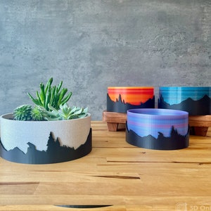 May include: Three different colored planters with a mountain range silhouette design. The planters are made of a 3D printed material and have a textured finish. The planters are sitting on a wooden surface.
