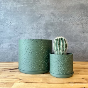 May include: Two green plant pots with a topographic design. One pot is larger and empty, while the other contains a small cactus. Both pots sit on matching saucers. The background is a gray wall, and the surface is a wooden table.