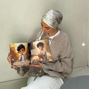 May include: A young person wearing a light brown sweater and a white headscarf with a gold and brown pattern is reading a children's book. The book features a cartoon illustration of a young boy with brown hair and a blue shirt. The text in the book reads "Meet Omar, a happy five year old, with a heart full of love and joy." and "Sometimes Omar struggles to find enough food to eat."