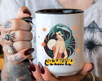 May include: White ceramic mug with a black rim and handle. The mug features a colorful illustration of a woman with a scorpion, stars, and the text "Scorpio".