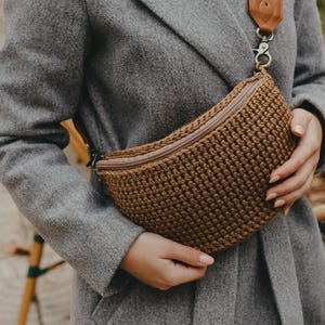 May include: A brown crochet fanny pack with a zipper closure and a patterned strap. The strap features a floral design with orange and black accents. The bag is held against a gray coat.