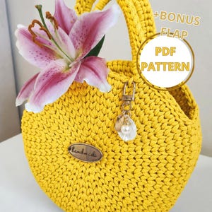 May include: A handmade, bright yellow crochet handbag with a rounded shape and a braided handle. A pink and white lily flower is tucked into the bag. The bag has a small metal charm and a tag that says "handmade". The image also includes text that says "+BONUS FLAP PDF PATTERN".