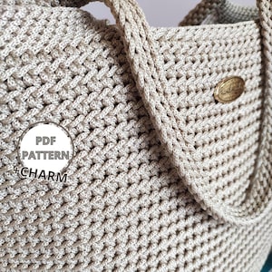 May include: Close-up of a beige crocheted tote bag with a textured weave pattern. The bag features braided handles and a small, oval-shaped gold tag. A circular graphic with the text "PDF PATTERN + CHARM" is visible.