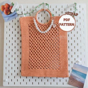 Könnte beinhalten: Eine pfirsichfarbene, gehäkelte Tragetasche mit netzartigem Design, rundem Griff und Perlenkette. Die Tasche hängt an einer weißen Lochwand. Das Bild enthält den Text "+STRAP PDF PATTERN".