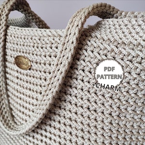 Op de afbeelding: Close-up van een beige gehaakte tote bag met gevlochten handvatten. De tas heeft een gestructureerd steekpatroon en een kleine, ronde, goudkleurige bedel. Een cirkelvormige afbeelding met de tekst "PDF PATTERN + CHARM" is zichtbaar.