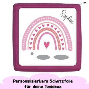 Könnte beinhalten: Eine quadratische, magenta Schutzhülle mit einem weißen Mittelteil, das ein rosa und pfirsichfarbenes Regenbogen-Design mit Herzen und dem Namen "Sophie" zeigt. Der Text "Personalisierbare Schutzfolie für deine Toniebox" befindet sich unten.