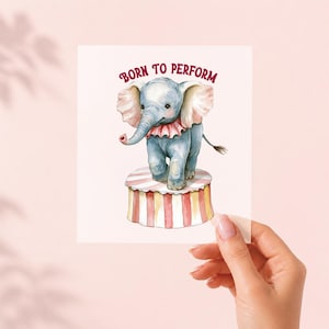 Puede incluir: Una tarjeta cuadrada con una ilustración en acuarela de un bebé elefante azul de pie sobre un pedestal de circo a rayas rojas y blancas. El elefante lleva un cuello con volantes y el texto BORN TO PERFORM aparece arqueado en rojo.