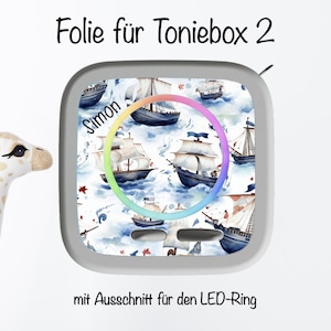 Könnte beinhalten: Dekorative Folie für eine Toniebox 2 mit einem nautischen Design, das Segelboote auf blau-weißem Hintergrund zeigt. Die Folie hat einen Ausschnitt für den LED-Ring und den Namen "Simon". Ein Giraffen-Spielzeug ist ebenfalls zu sehen.