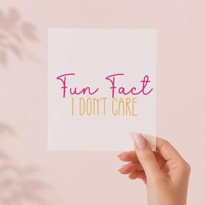 Può includere: Una carta rettangolare bianca con il testo Fun Fact in un carattere corsivo rosa, posizionato sopra il testo I DON'T CARE in un carattere sans-serif giallo. La carta è tenuta contro uno sfondo rosa chiaro con ombre morbide.
