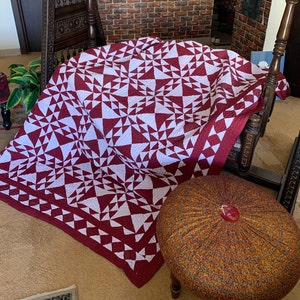 May include: A red and white quilt with a geometric pattern. The quilt is folded and draped over a wooden chair. A round, brown and gold patterned ottoman is in the foreground.