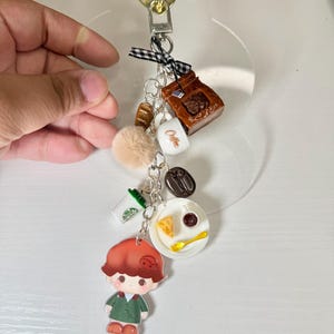 May include: A coffee-themed keychain with various charms, including a coffee bag, a coffee cup with "Coffee" text, a coffee bean, a pastry, a pom-pom, a small figure, and a plate with food. The keychain has a gold heart-shaped clasp.