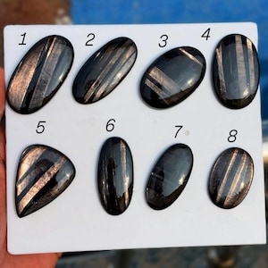 Puede incluir: Ocho piedras preciosas negras pulidas con rayas de color bronce metálico dispuestas en una tarjeta blanca. Cada piedra está numerada del uno al ocho, con superficies lisas y reflectantes y patrones lineales distintos en varias formas.