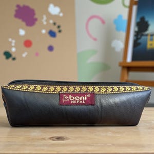 Op de afbeelding: Een rechthoekige etui van donkerbruin materiaal met een decoratieve goud-zwarte rand langs de rits. De etui heeft een rood label met de tekst "beni nepal". De etui ligt op een houten oppervlak.