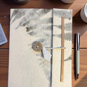 Op de afbeelding: Een handgemaakt notitieboek met een varenpatroon op de kaft. Het is vastgemaakt met een houten knoop en een bamboestok. Een zilveren pen ligt naast het notitieboek op een houten oppervlak. Kleine planten in potten staan op de achtergrond.