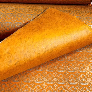 Könnte beinhalten: Nahaufnahme von gerolltem orangefarbenem Papier mit silbernem Blumenmuster. Das Papier hat eine strukturierte Oberfläche und eine gefaltete Ecke, die die Unterseite freilegt. Das Papier wird wahrscheinlich für Bastelarbeiten, Scrapbooking oder Geschenkverpackungen verwendet.