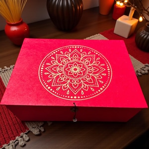 Può includere: Una scatola regalo rosso vivo con un design mandala dorato sul coperchio. La scatola è rettangolare con una superficie strutturata e una piccola chiusura a fermaglio. Il mandala è un motivo floreale dettagliato.