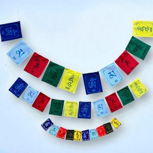 Könnte beinhalten: Bunte Gebetsfahnen in Blau, Rot, Grün und Gelb mit verschiedenen Symbolen und Texten. Die Fahnen sind an einer Schnur aufgereiht und in einem Bogen vor weißem Hintergrund angeordnet. Die Fahnen tragen die Schriftzüge "OM", "MA", "PADME" und "HUM".
