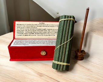 Peut inclure: Une boîte rouge avec un bouton doré contient un paquet de bâtonnets d'encens verts. La boîte porte le texte suivant : "KAPHA Mandala Arts & incense's Ayurvedic formula works with our other Ayurvedic product to help restore balance in the body, mind and air-prepared on the herbal smoke Inhalation procedure Your KAPHA Incense has been formulated to uplift, invigorate and stimulate Kapha dosha. We use our blends of Ayurvedic herbs, resin, spices and oil. Ingredients: Cymbopogen flexuous (Lemon grass) Leaf and oil, Base Cymbopogen nardus (Citronella), Myristica fragrance (Nutmeg), syrypum aromaticum. It contains 30 sticks with a burner. It burns 55 minutes approximate.  Un porte-encens en bois avec un bâtonnet d'encens brun se trouve à droite.