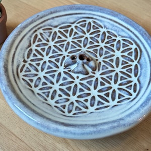 Könnte beinhalten: Ein Keramik-Räucherstäbchenhalter in Hellblau und Braun. Der Halter hat ein detailliertes geometrisches Muster in der Mitte und ein kleines Loch für Räucherstäbchen. Der runde Halter hat einen erhöhten Rand. Durchmesser ca. 15 cm.
