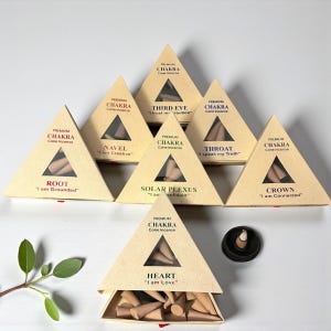 Könnte beinhalten: Eine Auswahl an Chakra-Kegel-Räucherstäbchen-Sets in dreieckigen Schachteln. Jede Schachtel ist mit einem Chakra-Namen und einer Affirmation beschriftet, wie z. B. "Root - I am Grounded" und "Heart - I am Love". Ein kleiner schwarzer Räucherstäbchenhalter ist ebenfalls vorhanden.