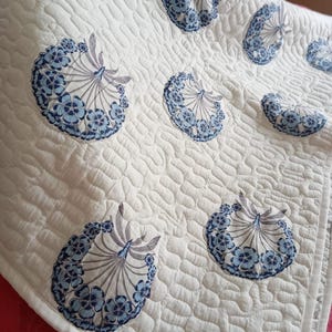 Peut inclure: Une couverture matelassée blanche avec un motif répété de motifs floraux bleus et blancs en forme d'éventail. La couverture a une surface texturée et une bordure blanche. Le design est de style vintage.