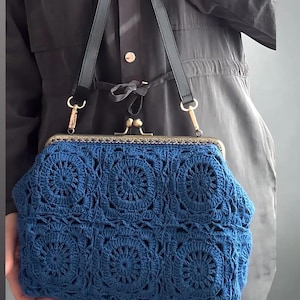 Könnte beinhalten: Eine blaue Häkeltasche mit Metallverschluss und schwarzem Schulterriemen. Die Tasche hat ein detailliertes Blumenmuster und einen goldfarbenen Rahmen. Die Tasche wird von einer Person gehalten, die eine dunkelgraue Jacke trägt.