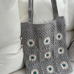 Könnte beinhalten: Graue gehäkelte Tragetasche mit floralem Design. Die Tasche hat ein quadratisches Muster mit weißen und teal-farbenen Blumenmotiven. Die Tragetasche hat zwei lange Träger zum Tragen und ist aus weichem, strukturiertem Garn gefertigt.