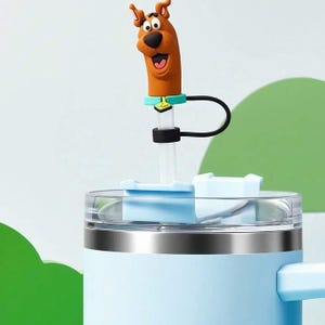 Könnte beinhalten: Ein hellblauer Becher mit einem silbernen Band und einem transparenten Deckel. Der Becher hat einen Strohhalmaufsatz mit der Cartoonfigur Scooby-Doo. Der Strohhalm ist schwarz und weiß, und der Aufsatz ist braun, schwarz und orange.