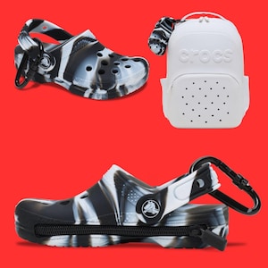 May include: A set of Crocs shoes and accessories against a red background. One shoe is black and white marbled, with a black zipper and a black clip. A white backpack with the Crocs logo and a black and white marbled charm is also shown.