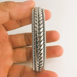Op de afbeelding: Een zilverkleurige armband met een gevlochten ontwerp in het midden en een touwachtige rand. De armband is gemaakt van glanzend metaal en ziet er nieuw uit.