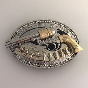 May include: Oval-shaped silver and gold belt buckle featuring a revolver design. The buckle has a silver rope-like border and a detailed floral pattern. The revolver is silver and gold, with the text "COLT 45" visible.