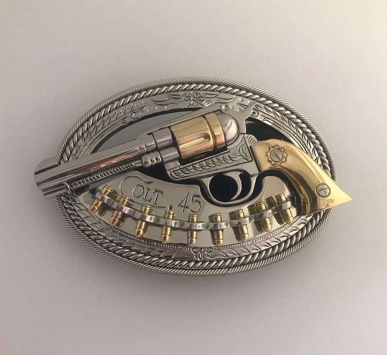 May include: Oval-shaped silver and gold belt buckle featuring a revolver design. The buckle has a silver rope-like border and a detailed floral pattern. The revolver is silver and gold, with the text "COLT 45" visible.
