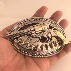 May include: Silver and gold-toned metal belt buckle featuring a revolver design with the text "COLT .45". The buckle has an oval shape with a rope-like border and decorative floral elements.