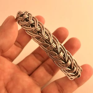 Peut inclure: Un bracelet argenté avec un motif tressé. Le bracelet est composé de plusieurs brins de métal entrelacés pour créer un motif texturé. Le bracelet est tenu dans une main.