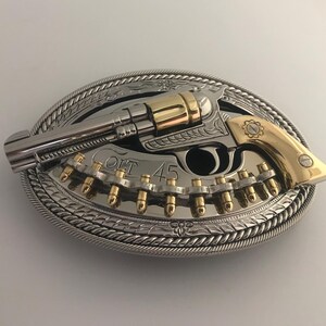 May include: An oval-shaped silver and gold belt buckle featuring a detailed revolver design. The gun is silver with gold accents, and the buckle includes the text "COLT 45". The buckle has a rope-like border.